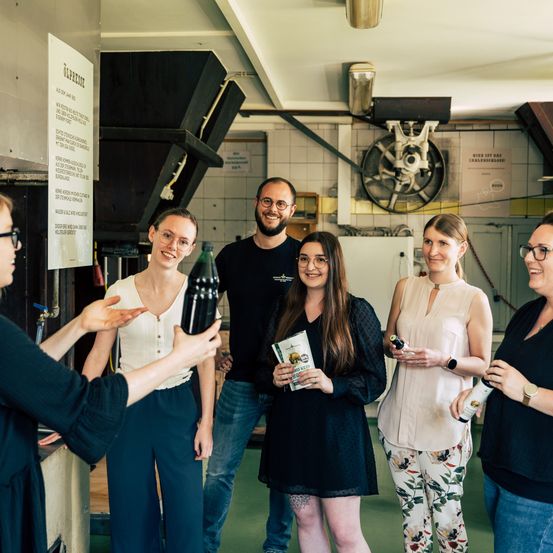Eine Gruppe von Menschen versammelt sich in einer industriellen Umgebung, lächelt und posiert für ein Foto. Eine Frau hält eine Flasche und ein Buch, während eine andere eine Flasche hält. Ein Mann mit Brille und Bart steht in der Mitte. Hinter ihnen sind Geräte und eine Maschine zu sehen.