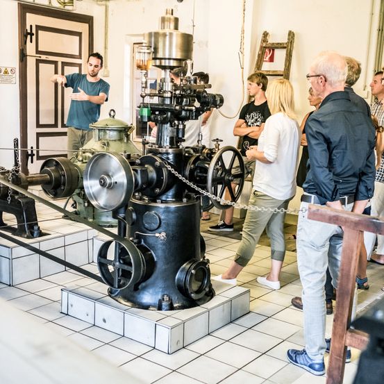 Ein Mann erklärt einer Gruppe von Menschen in einem Museum eine alte mechanische Pumpe.