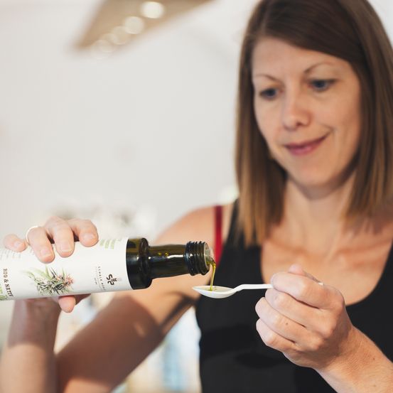Eine Frau gießt Öl aus einer Flasche in einen weißen Löffel, lächelt und hält ihn in ihrer rechten Hand.