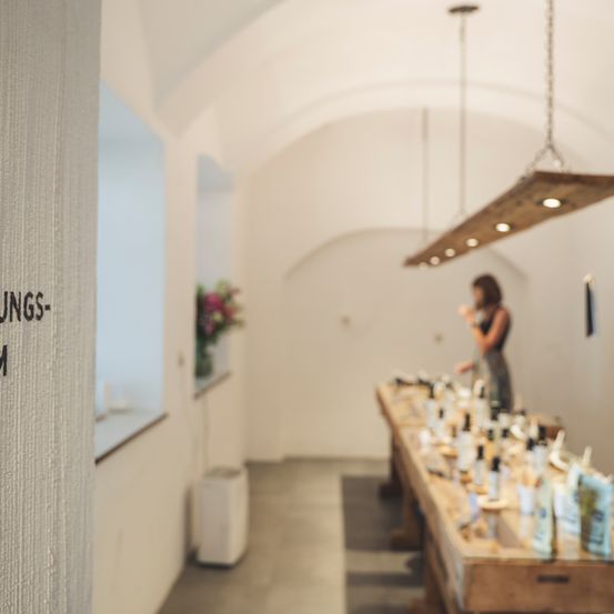 Eine Frau steht in einem Raum mit weißen Wänden und einem gewölbten Dach. Es gibt einen langen Tisch mit Flaschen und Gegenständen darauf. Eine Pflanze steht im Fenster.
