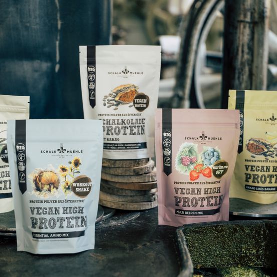Mehrere vegane Proteinpulver-Beutel von Schalk Muehle, einschließlich eines mit der Aufschrift 'Workout Shake', sind auf einem Tisch angeordnet, vor einem Hintergrund aus gestapelten Holzfässern.