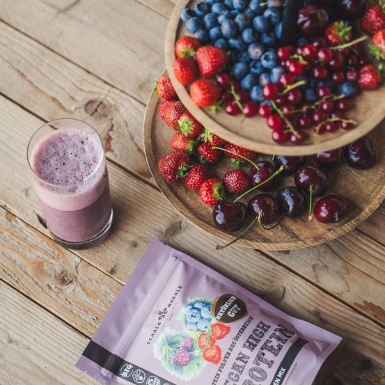 Ein Smoothie in einem Glas mit einer Schüssel voller Beeren, darunter Blaubeeren, Erdbeeren und Kirschen, auf einem Holztisch. Ein Beutel veganes Proteinpulver ist in der Nähe.