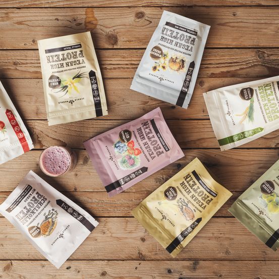 Verschiedene Tüten mit unterschiedlichen Proteinpulvern sind auf einer Holzoberfläche ausgestellt, einschließlich Geschmacksrichtungen wie Blaubeere, Vanille und Schokolade. Jede Tüte ist mit ihrem Geschmack und ihren Nährwertangaben beschriftet.