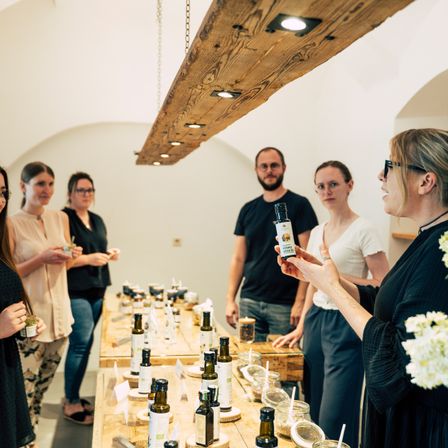 Eine Gruppe von Menschen versammelt sich um einen Tisch mit verschiedenen Ölflaschen, angeführt von einer Frau, die eine Flasche hält, mit einer Holzdecke und einer weißen Wand im Hintergrund.