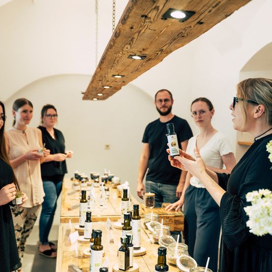 Eine Gruppe von Menschen versammelt sich um einen Tisch mit verschiedenen Ölflaschen, angeführt von einer Frau, die eine Flasche hält, mit einer Holzdecke und einer weißen Wand im Hintergrund.