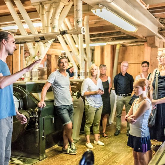 Eine Gruppe von Menschen, darunter ein Mann in einem blauen T-Shirt und ein anderer in einem grauen T-Shirt, versammelt sich in einer Fabrik. Sie scheinen einem Mann zuzuhören, der etwas über die Maschinen erklärt. Der Raum hat Holzwände und Rohre.