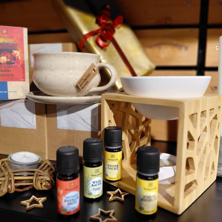 Eine Holzbox enthält eine Schüssel, eine Tasse und einen Untertasse. Verschiedene Flaschen mit ätherischen Ölen sind um die Schüssel herum platziert. Ein Buch befindet sich neben der Tasse, und eine Geschenkbox mit einem roten Band ist hinter der Tasse.