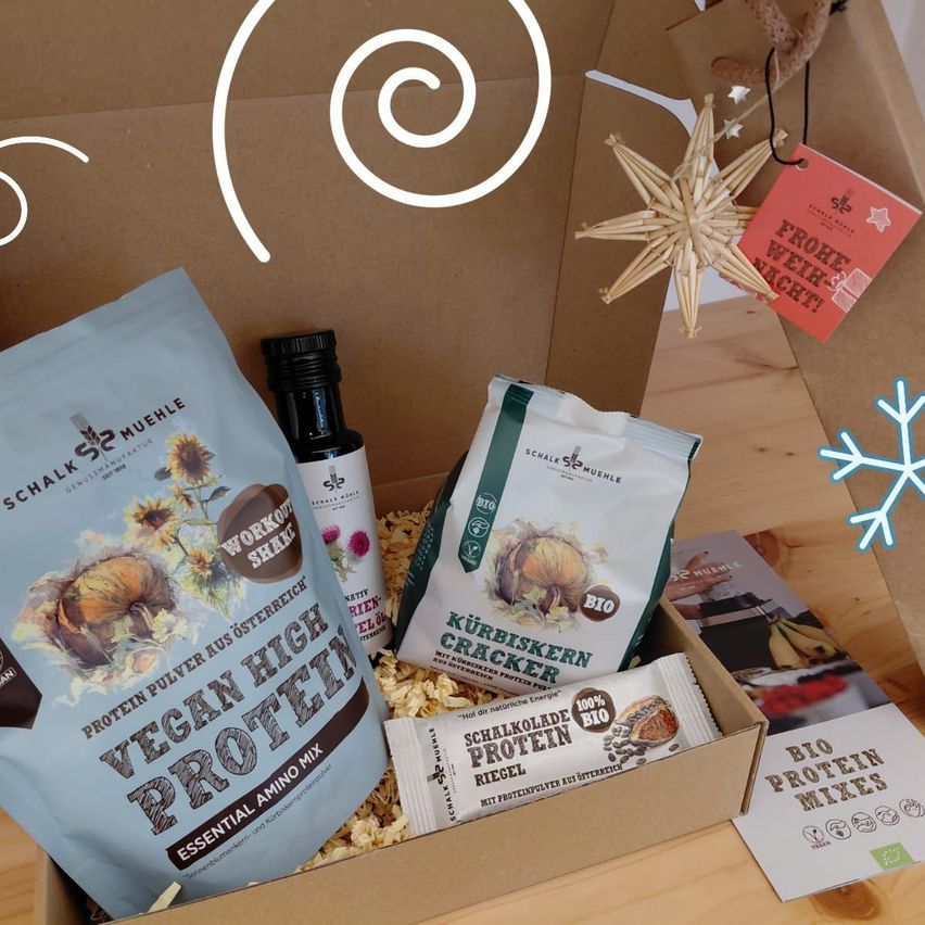 Eine Box gefüllt mit veganen Produkten. Eine Flasche Öl, ein Beutel mit Protein und ein Beutel mit Crackern. Außerdem eine Weihnachtskarte und eine Schneeflockendekoration.