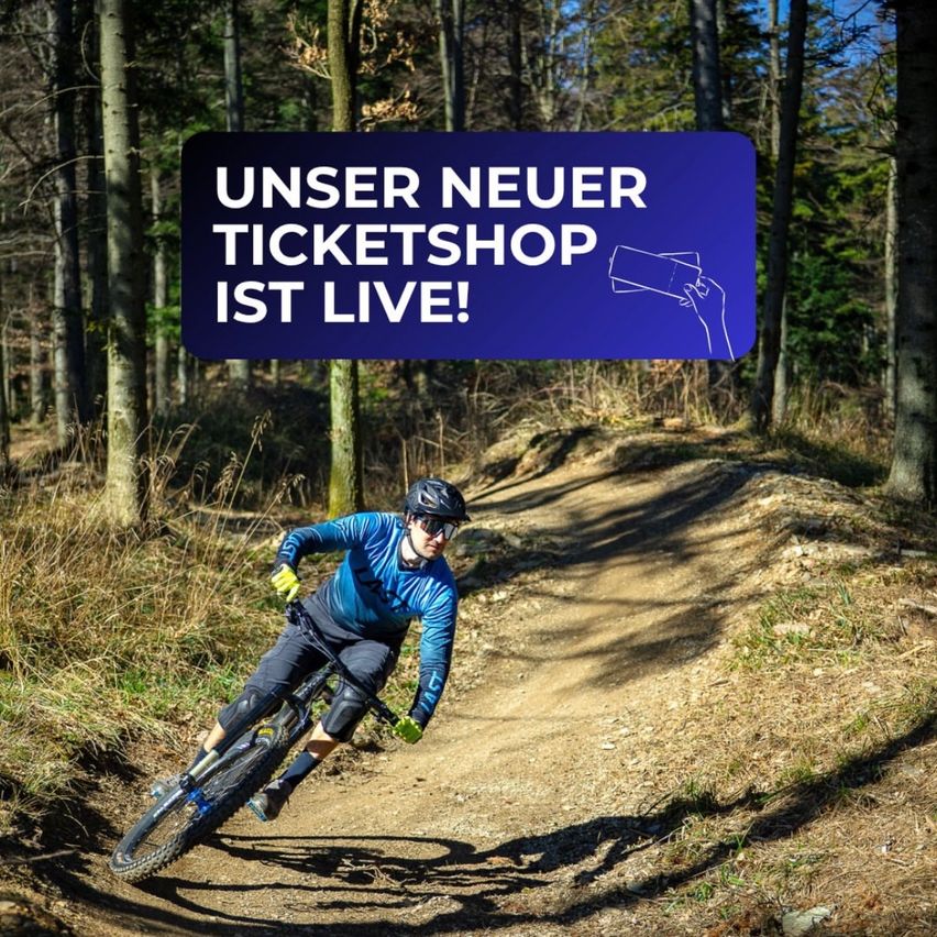 Ein Mann fährt ein Mountainbike auf einem Weg in einem Wald. Ein überlagerter Text lautet 'Unser neuer Ticketshop ist live!' Der Mann trägt einen Helm und Handschuhe.