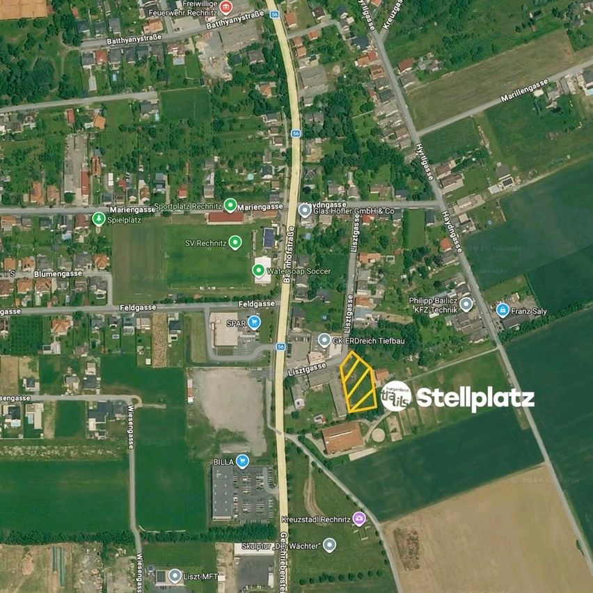 Eine Luftaufnahme einer kleinen Stadt mit Wohngebieten, grünen Feldern und einem Straßennetz. Bemerkenswerte Punkte sind ein Sportplatz, eine Fabrik und ein gekennzeichnetes Grundstück 'Stellplatz'.