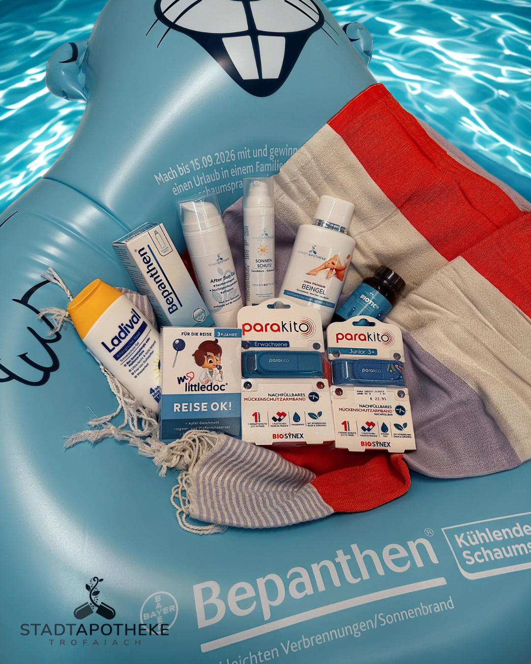 Ein blauer aufblasbarer Schwimmring zeigt verschiedene Hautpflegeprodukte wie Sonnencreme, After-Sun-Lotion und Babypflegeprodukte. Der Schwimmring ist teilweise im Wasser eingetaucht, ein Handtuch und ein Handtuchständer sind sichtbar.