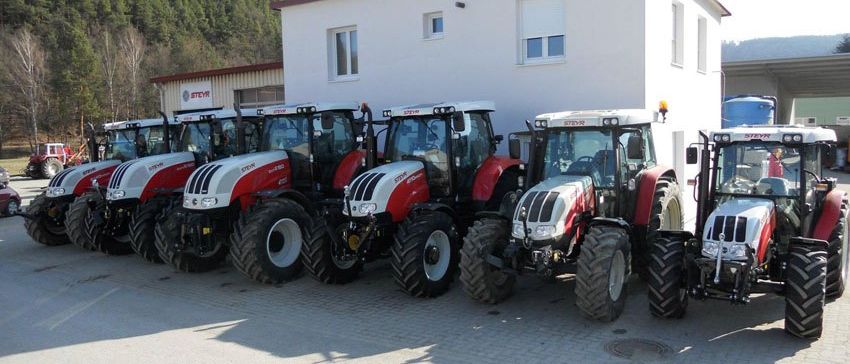 Mehrere rote und weiße Traktoren sind vor einem weißen Gebäude mit Fenstern und einem Logo an der Seite geparkt. Hinter den Traktoren befindet sich ein Berg mit Bäumen und einem blauen Fass.