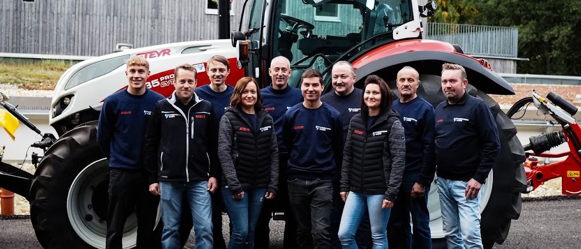 Eine Gruppe von Personen in gleichen blauen und weißen Jacken posiert vor einem großen roten und weißen Traktor. Sie sind draußen auf einem Parkplatz mit einem Gebäude und Bäumen im Hintergrund.