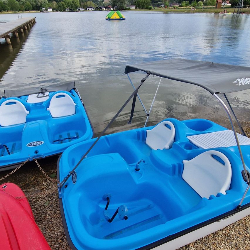 Zwei blaue Tretboote sind am Ufer eines Sees verankert, eines mit einem Sonnendach. Ein roter Kajak ist in der Nähe. Ein bunter Schwimmkörper ist in der Ferne. Bäume und Gebäude sind im Hintergrund zu sehen.