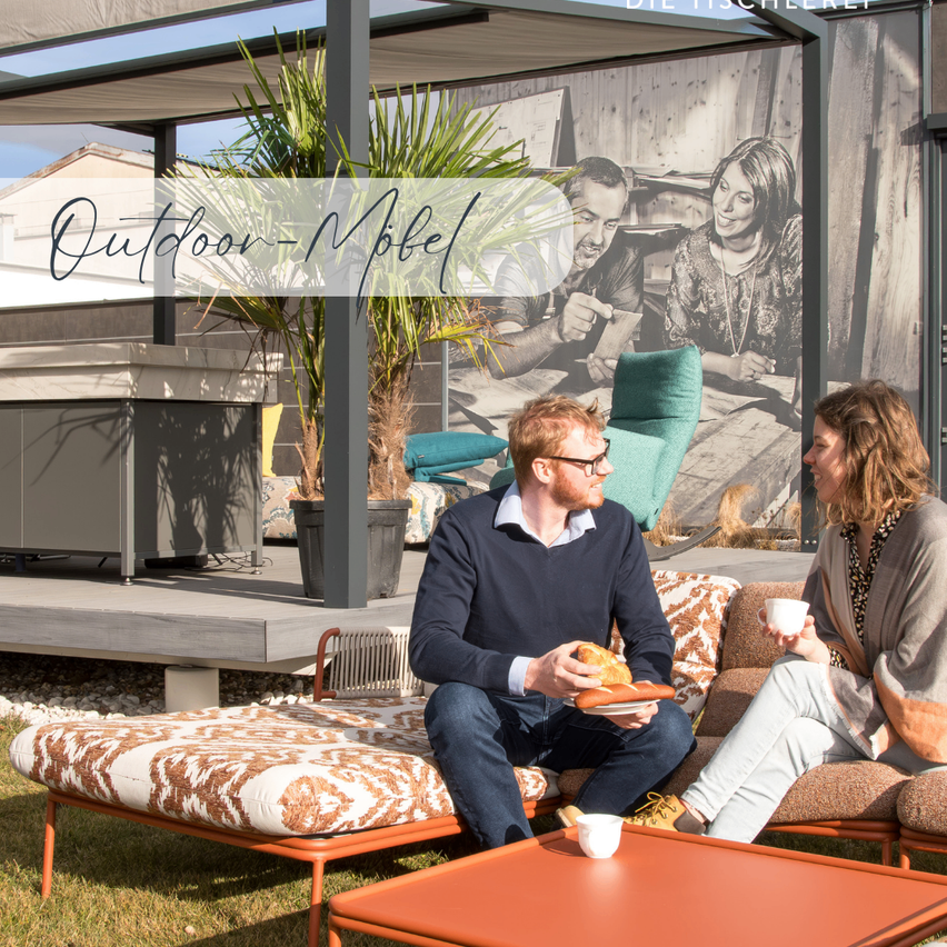 Two people sit on a couch in an outdoor setting, holding cups. A large image is behind them, and a plant is in front of them.