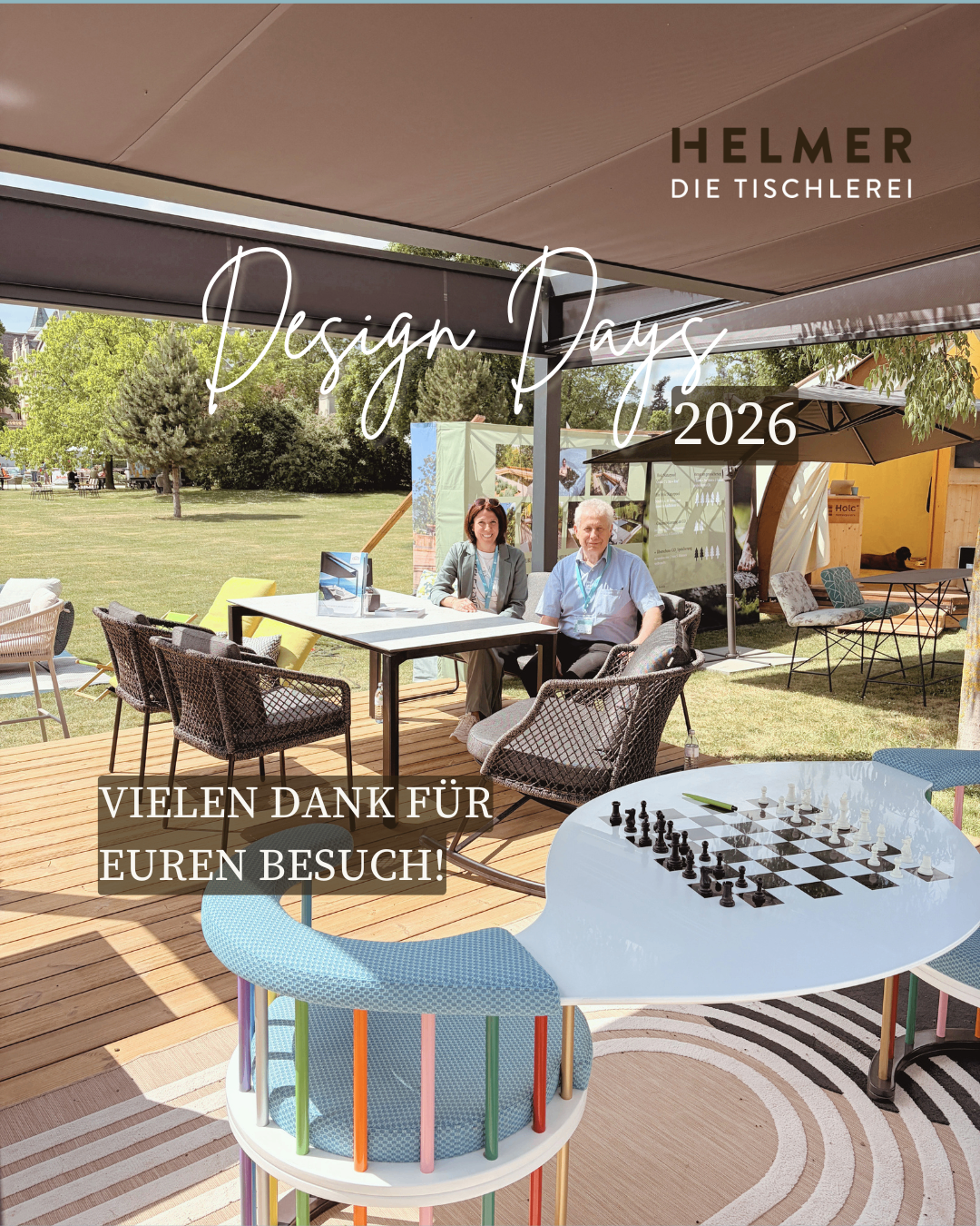 Zwei Personen sitzen an einem Tisch auf einer Terrasse mit einem Schachspiel, unter einem Baldachin mit dem Text Helmer Design Days 2026. Die Umgebung ist im Freien, mit Bäumen und einem Rasen im Hintergrund.
