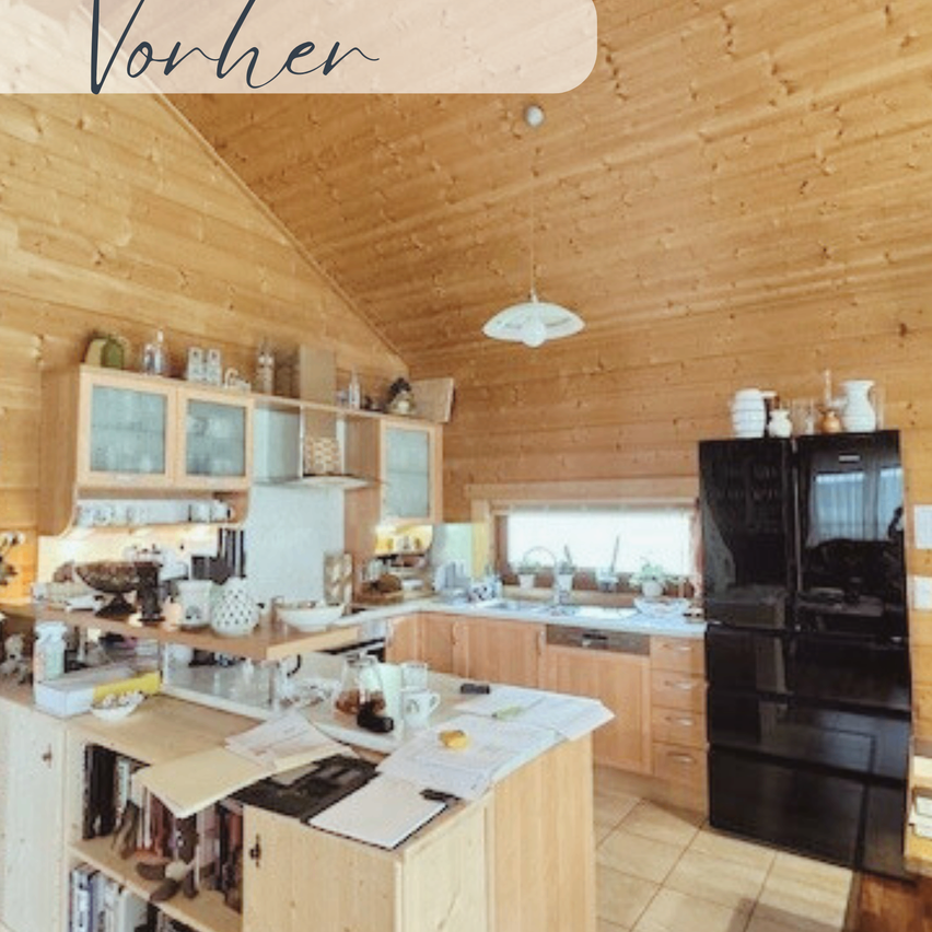 Eine gemütliche Küche mit Holzwänden und Schränken. Ein Kühlschrank und ein Schrank befinden sich auf der rechten Seite. Links steht ein Schreibtisch mit Büchern, Papieren und einem Wasserkocher. Eine Lampe hängt von der Decke.