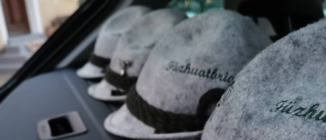 Mehrere graue Fedoras mit schwarzen Bändern und deutschem Text sind in einem Fahrzeug gestapelt. Ein Haus ist im Hintergrund sichtbar.