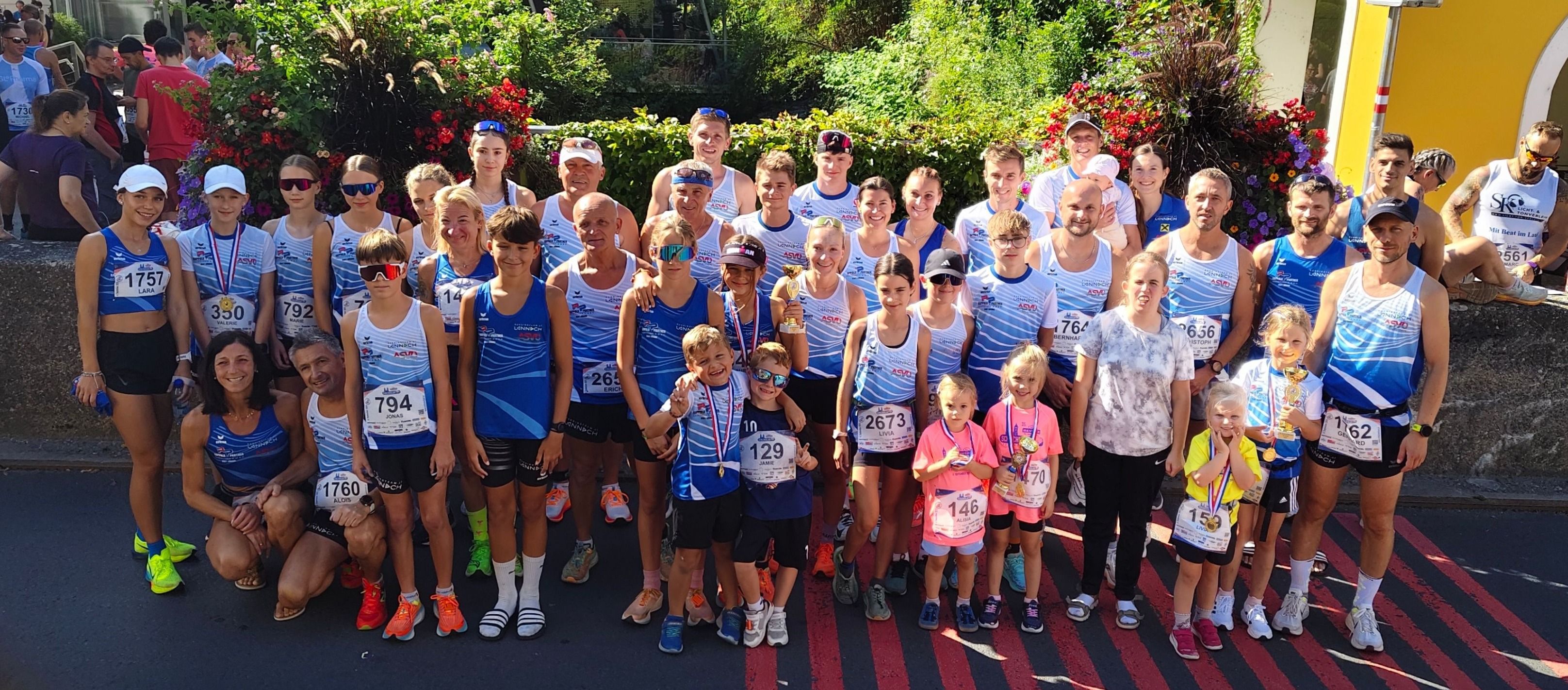 Eine Gruppe von Läufern, sowohl Erwachsene als auch Kinder, stehen in einer Reihe, tragen blaue und weiße Uniformen, Medaillen und Turnschuhe. Sie lächeln und posieren für ein Foto. Sie stehen auf einer Straße mit einer roten Linie.