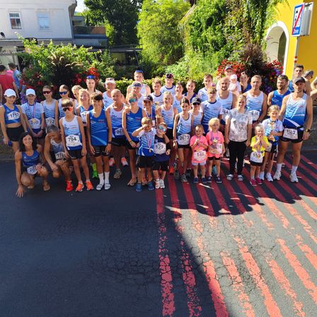 Eine Gruppe von Menschen in Laufausruestung und mit Startnummern posiert fuer ein Foto auf der Strasse, mit Pflanzen und Gebaeuden im Hintergrund.