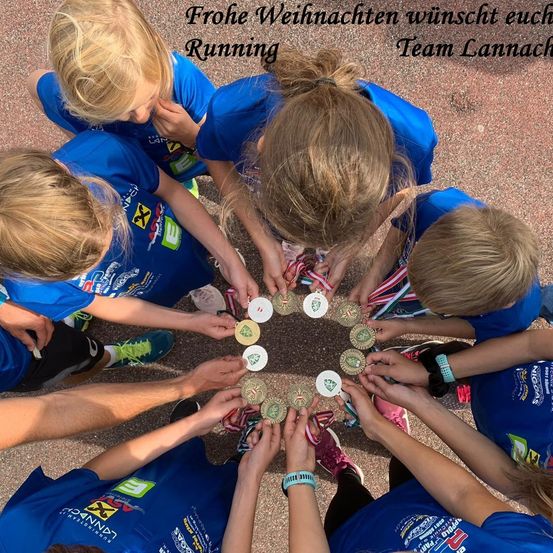 Gruppe von Kindern in blauen Trikots mit Medaillen versammelt sich im Kreis, mit einer festlichen Weihnachtsbotschaft von Team Lannach über ihnen.