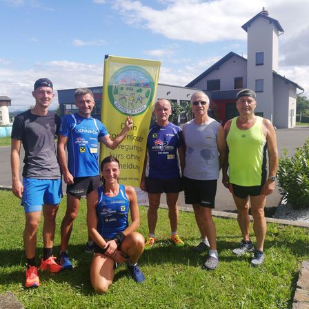 Eine Gruppe von Menschen in Sportbekleidung steht auf einem Rasenfeld. Eine Frau kniet vorne, und ein Mann hält ein gelbes Banner mit einer Botschaft. Hinter ihnen steht ein weißes Gebäude.