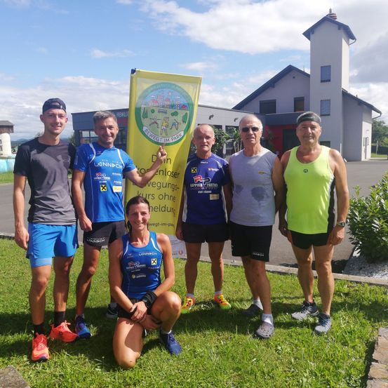 Eine Gruppe von Menschen in Sportbekleidung steht auf einem Rasenfeld. Eine Frau kniet vorne, und ein Mann hält ein gelbes Banner mit einer Botschaft. Hinter ihnen steht ein weißes Gebäude.