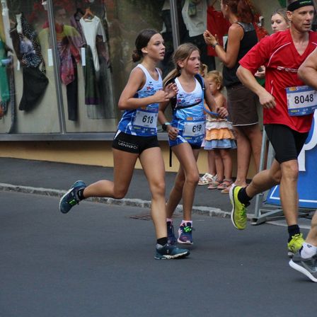 Eine Läuferin mit der Nummer 618 führt einen Marathon an, dicht gefolgt von einer Läuferin mit der Nummer 619, beide in Tanktops. Zuschauer, darunter Kinder, beobachten hinter einer Schaufensterscheibe.