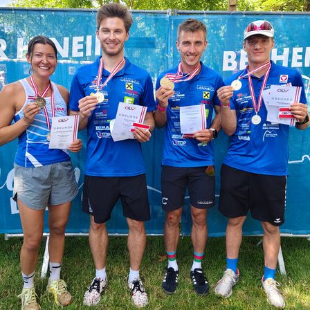 Vier Athleten stehen lächelnd zusammen, tragen blaue Shirts, schwarze Shorts und Turnschuhe. Sie halten Goldmedaillen und Urkunden. Der Hintergrund ist ein blauer Banner mit weißem Text.