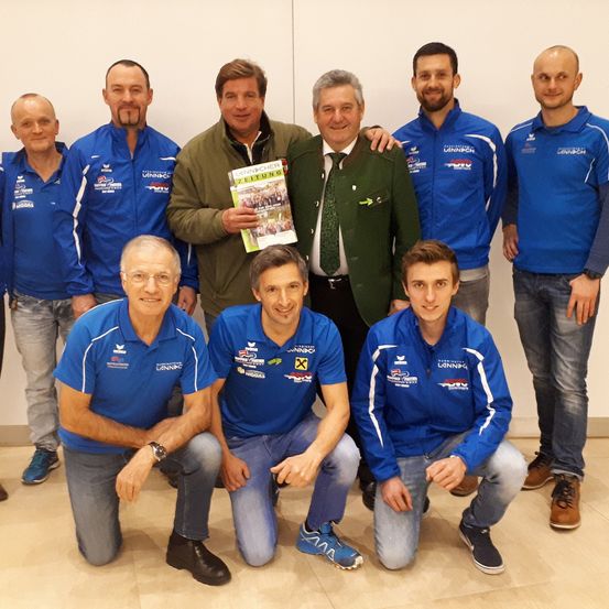 Eine Gruppe von Männern in blauen Jacken und Jeans posiert für ein Foto. Ein Mann in einer grünen Jacke hält ein Buch. Sie stehen vor einer weißen Wand.