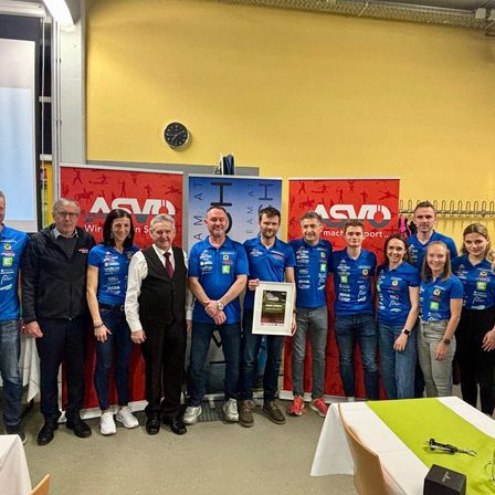 Eine Gruppe von Menschen in blauen Trikots und Turnschuhen steht vor Bannern und einem Tisch. Ein Mann hält einen gerahmten Preis.