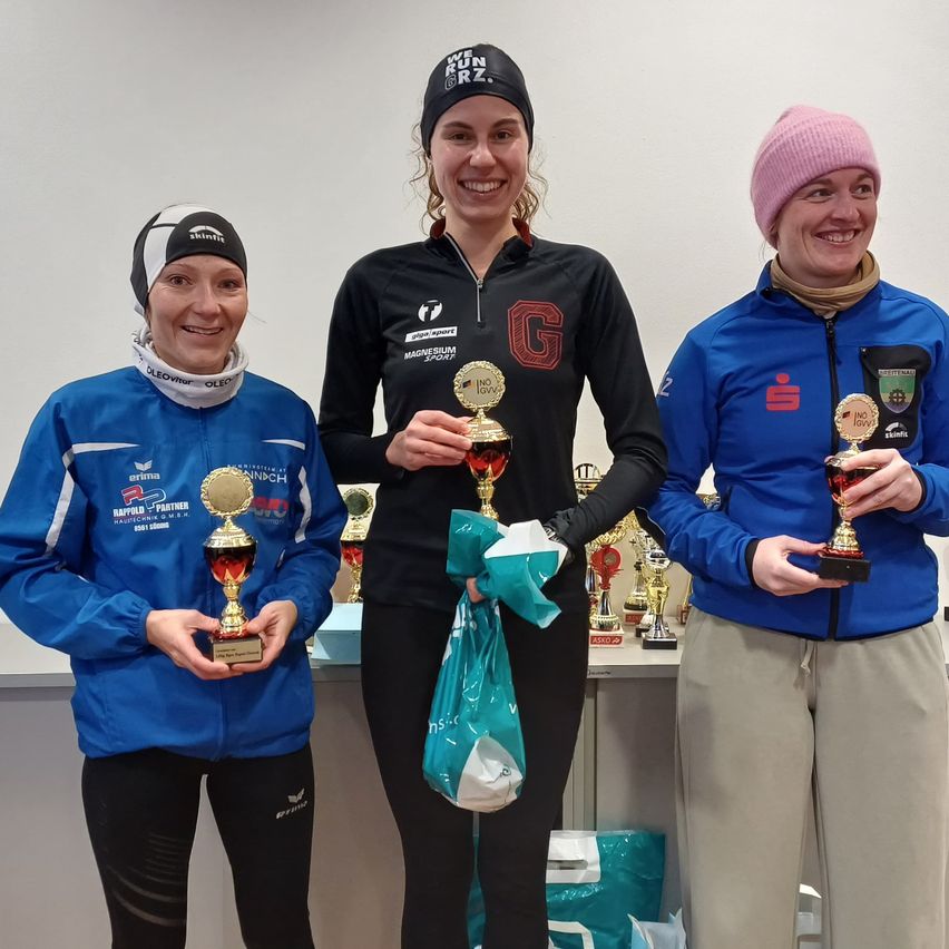 Drei Frauen stehen lächelnd nebeneinander und halten Pokale. Die Frau in der Mitte trägt eine schwarze Jacke und eine Kopfband, während die anderen blaue Jacken tragen. Sie stehen vor einer weißen Wand, mit Pokalen auf einem Tisch dahinter.