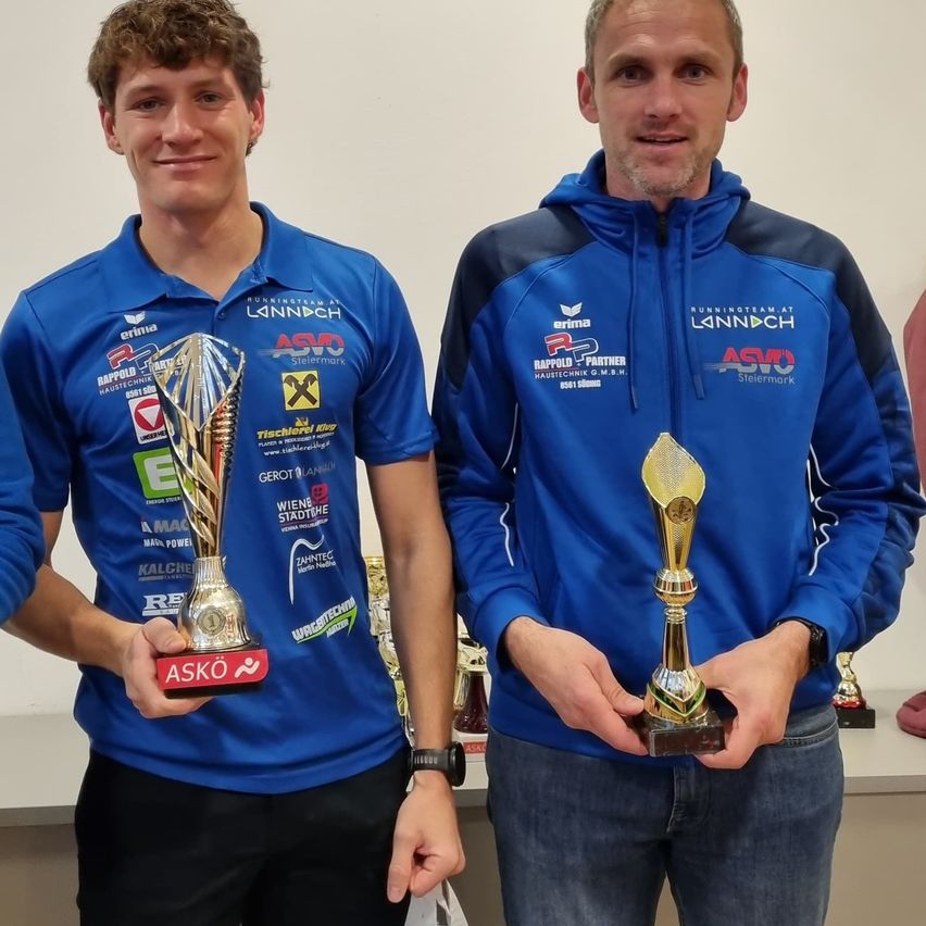 Zwei Männer in blauen Hemden stehen nebeneinander und halten Pokale. Sie lächeln und posieren für ein Foto.