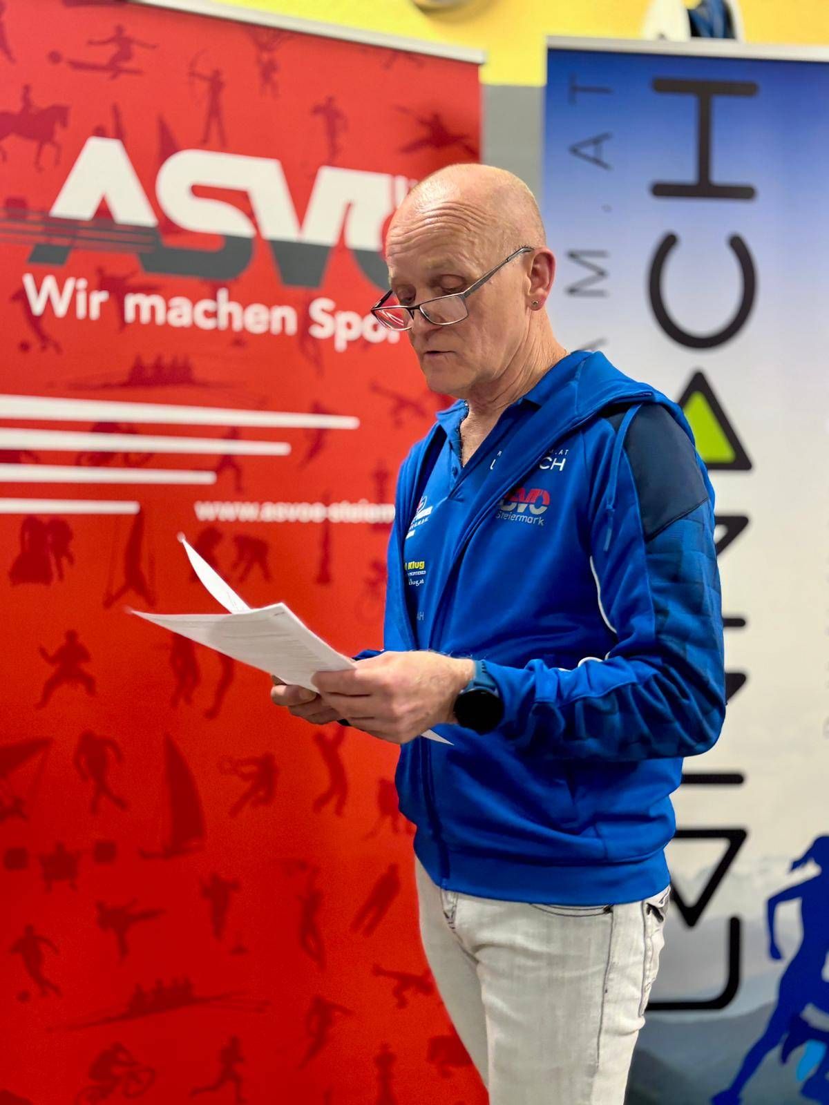 Ein älterer Mann in einem blauen Jackett liest ein Papier vor einer roten Wand mit dem Text 'Wir machen Sport'. Er trägt eine Brille.