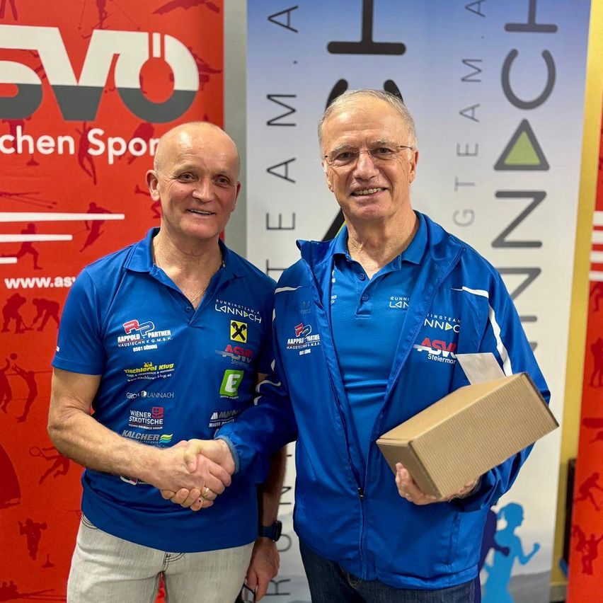 Zwei Männer in blauen Jacken stehen nebeneinander und lächeln. Ein Mann schüttelt die Hand des anderen. Sie halten eine braune Box. Hinter ihnen befinden sich zwei Banner.