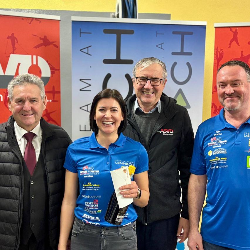 Vier Personen stehen gemeinsam vor einem Werbebanner. Eine Frau hält ein Papier, und die Männer lächeln.