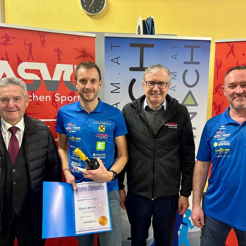 Vier Männer stehen nebeneinander vor einer Wand mit Bannern. Der Mann in der Mitte hält eine Flasche und ein Zertifikat. Sie alle lächeln.