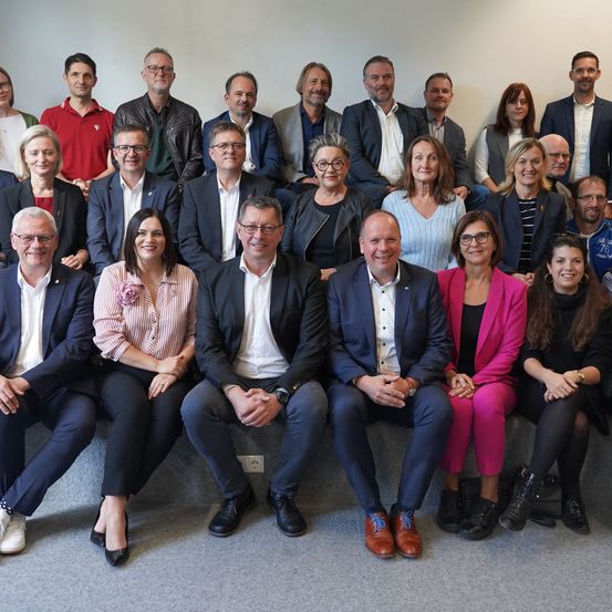 Ein Gruppenfoto von Fachleuten, gekleidet in formeller Kleidung, die zusammen auf einer Bank sitzen. Sie scheinen für eine Kamera in einem gut beleuchteten Raum zu posieren.