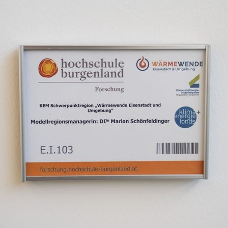 Ein weißes Schild mit orangefarbenen Akzenten zeigt Logos und Text auf Deutsch. Es identifiziert die Institution als 'Hochschule Burgenland' und listet einen Forschungsschwerpunkt auf 'Wärmewende Eisenstadt und Umgebung'. Das Schild enthält einen Barcode, den Namen des Modellregionsmanagers und eine Website-Adresse.