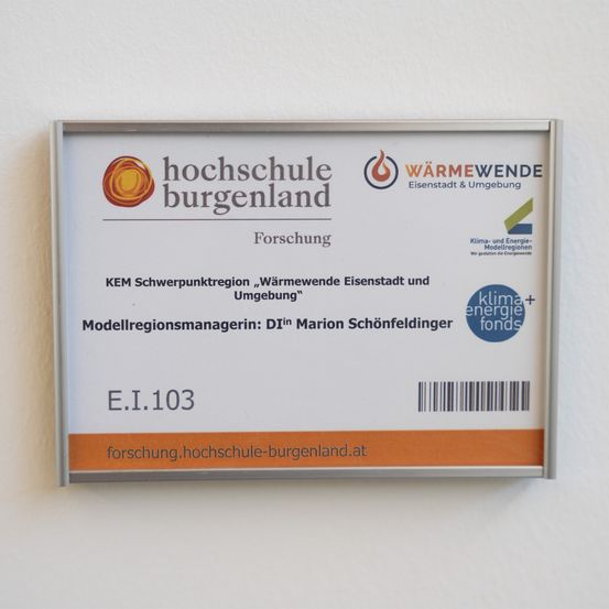 Ein weißes Schild mit orangefarbenen Akzenten zeigt Logos und Text auf Deutsch. Es identifiziert die Institution als 'Hochschule Burgenland' und listet einen Forschungsschwerpunkt auf 'Wärmewende Eisenstadt und Umgebung'. Das Schild enthält einen Barcode, den Namen des Modellregionsmanagers und eine Website-Adresse.