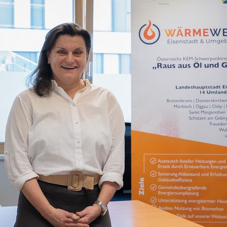 Eine Frau in einem weißen Hemd und einer grauen Hose steht vor einem Tisch mit Blumen. Hinter ihr befindet sich ein Plakat mit dem Text 'WärmeWeisenstadt' und Informationen zu einer Veranstaltung.