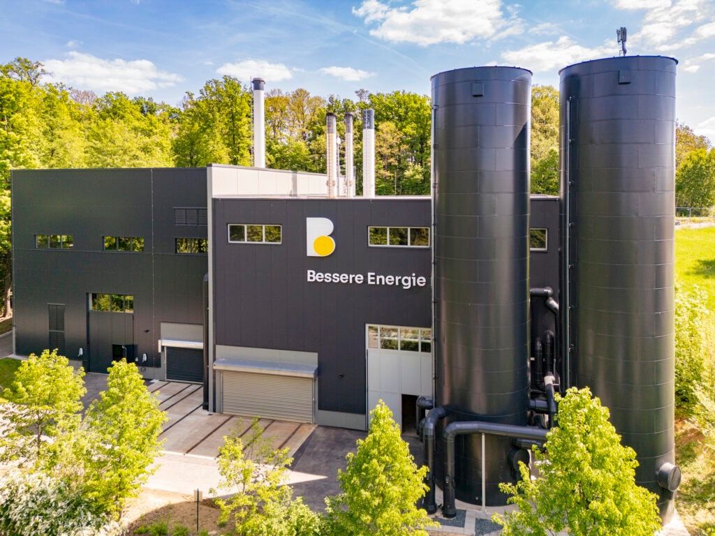 Eine moderne Industrieanlage mit einem großen 'B'-Logo und den Worten 'Bessere Energie' an der Wand, umgeben von Bäumen und einem klaren Himmel.