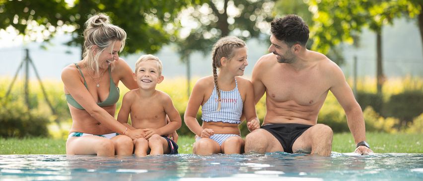 Eine Familie mit zwei Kindern, einem Jungen und einem Mädchen, lächelt, während sie am Rand eines Pools sitzen. Der Junge trägt ein Armband und das Mädchen trägt einen Badeanzug mit dem Namen Tommy.
