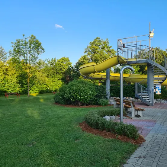 Eine gelbe Wasserrutsche mit Treppen, in einem Park gelegen. Die Rutsche ist von einem gepflegten grünen Rasen und verschiedenen Pflanzen umgeben. Bäume befinden sich im Hintergrund.