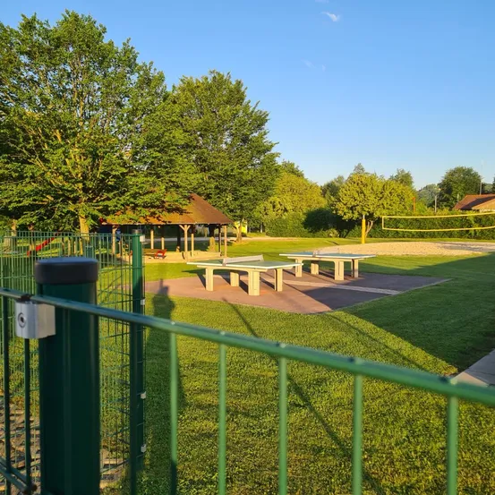 Ein Park mit Spielplatz, Pavillon und zwei Tischtennisplatten, umgeben von saftigem grünem Rasen. Bäume und Büsche sind in der Umgebung.