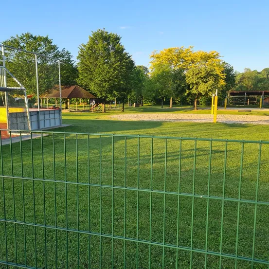 Ein gut gepflegter Park mit Spielplatz, Pavillon und Sportgeräten. Der grüne Zaun umschließt einen Rasenbereich. Bäume säumen den Park, und der Himmel ist klar.