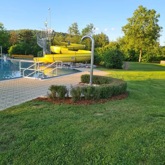 Ein Wasserpark mit einem gelben Wasserrutsche neben einem Pool. Die Rutsche führt zum Wasser. Es gibt Handläufe und Treppen um den Pool. Das Gelände ist von Bäumen und Gras umgeben.