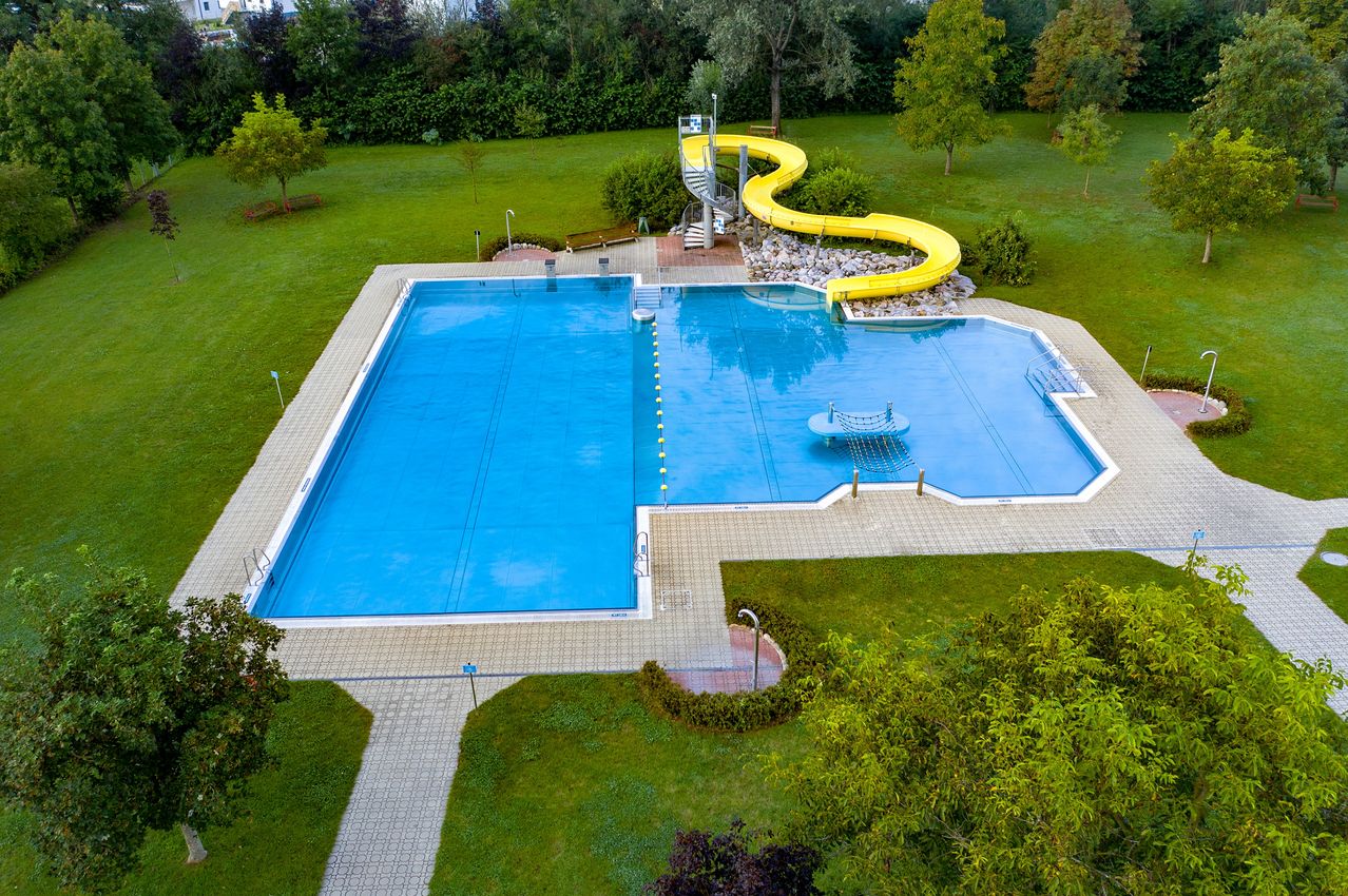 Ein Luftbild eines großen Schwimmbeckens mit einer gelben Wasserrutsche. Das Becken ist rechteckig und mit klarem blauem Wasser gefüllt. Es gibt Bäume und Büsche rund um das Becken.