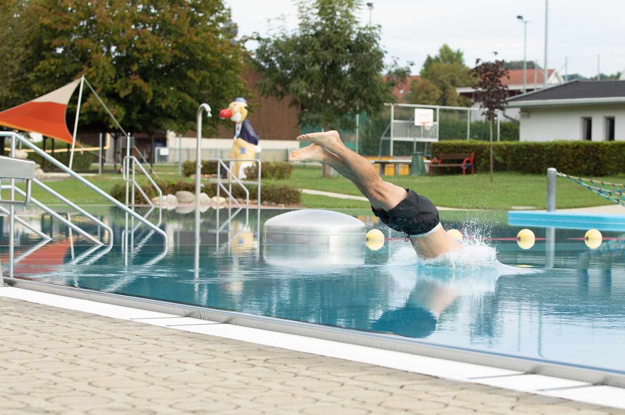 A man dives into an outdoor pool with a yellow ball floating nearby, surrounded by trees and a playground.
