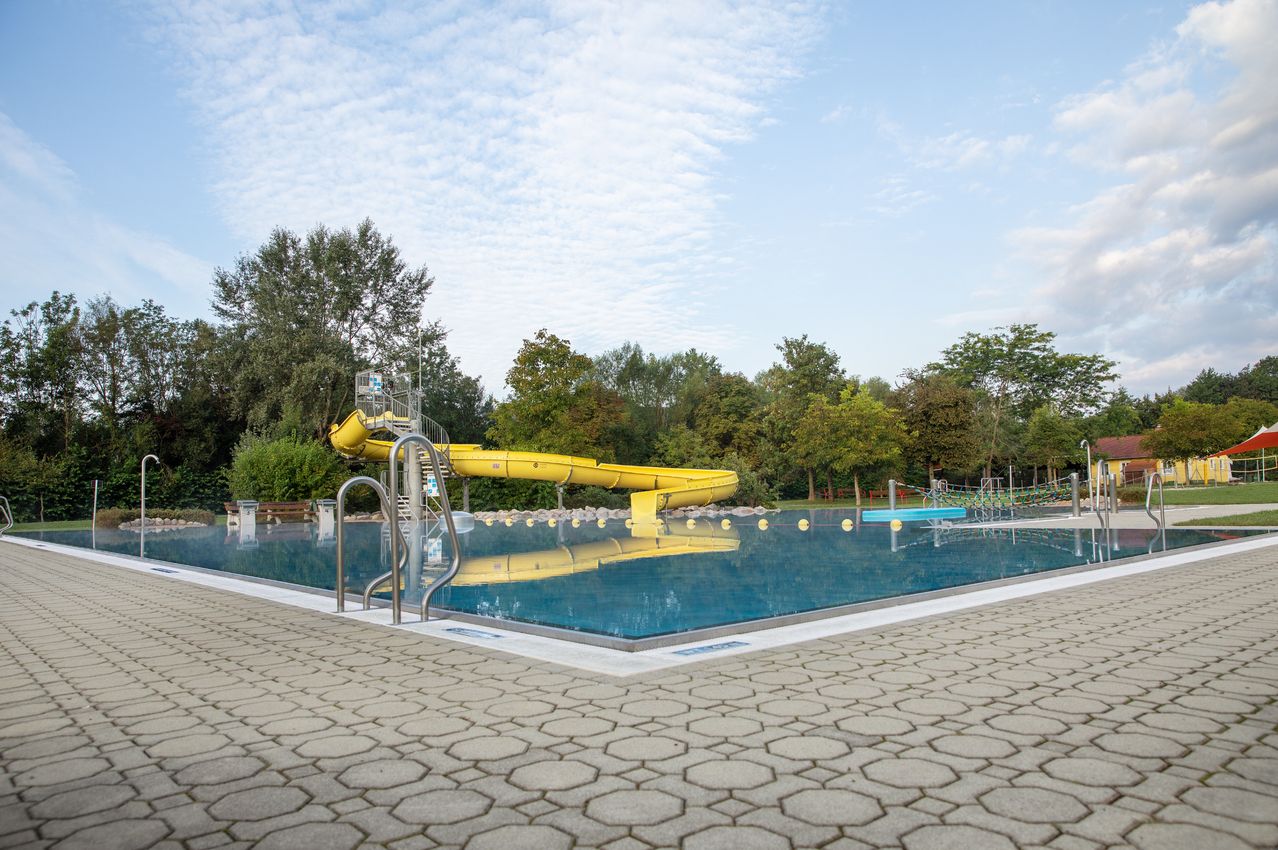 Ein Freibad mit einem gelben Wasserrutsche am Rand, umgeben von Bäumen und Pflanzen unter einem blauen Himmel.
